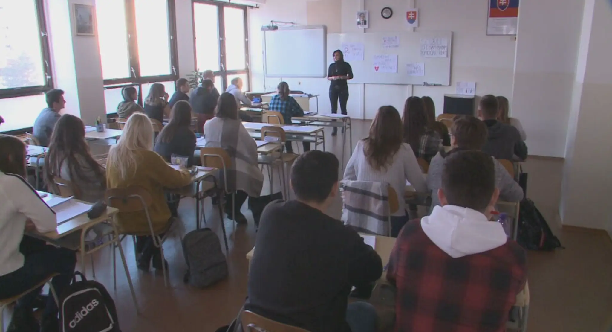 Na snímke sú študenti strednej školy, sedia v laviciach a počúvajú učiteľku, ktorá stojí pred tabuľou.