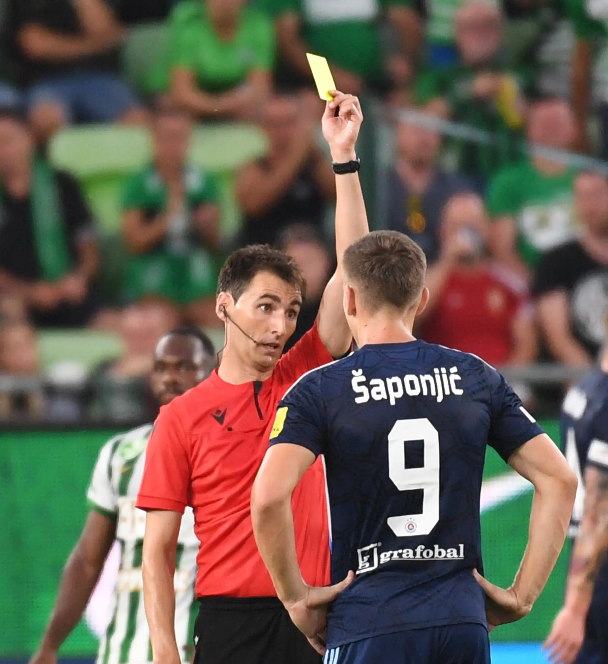 Hlavný rozhodca Ricardo de Burgos udeľuje žltú kartu hráčovi Slovana Ivanovi Šaponjičovi počas úvodného zápasu 2. predkola LM na pôde Ferencvárosu.
