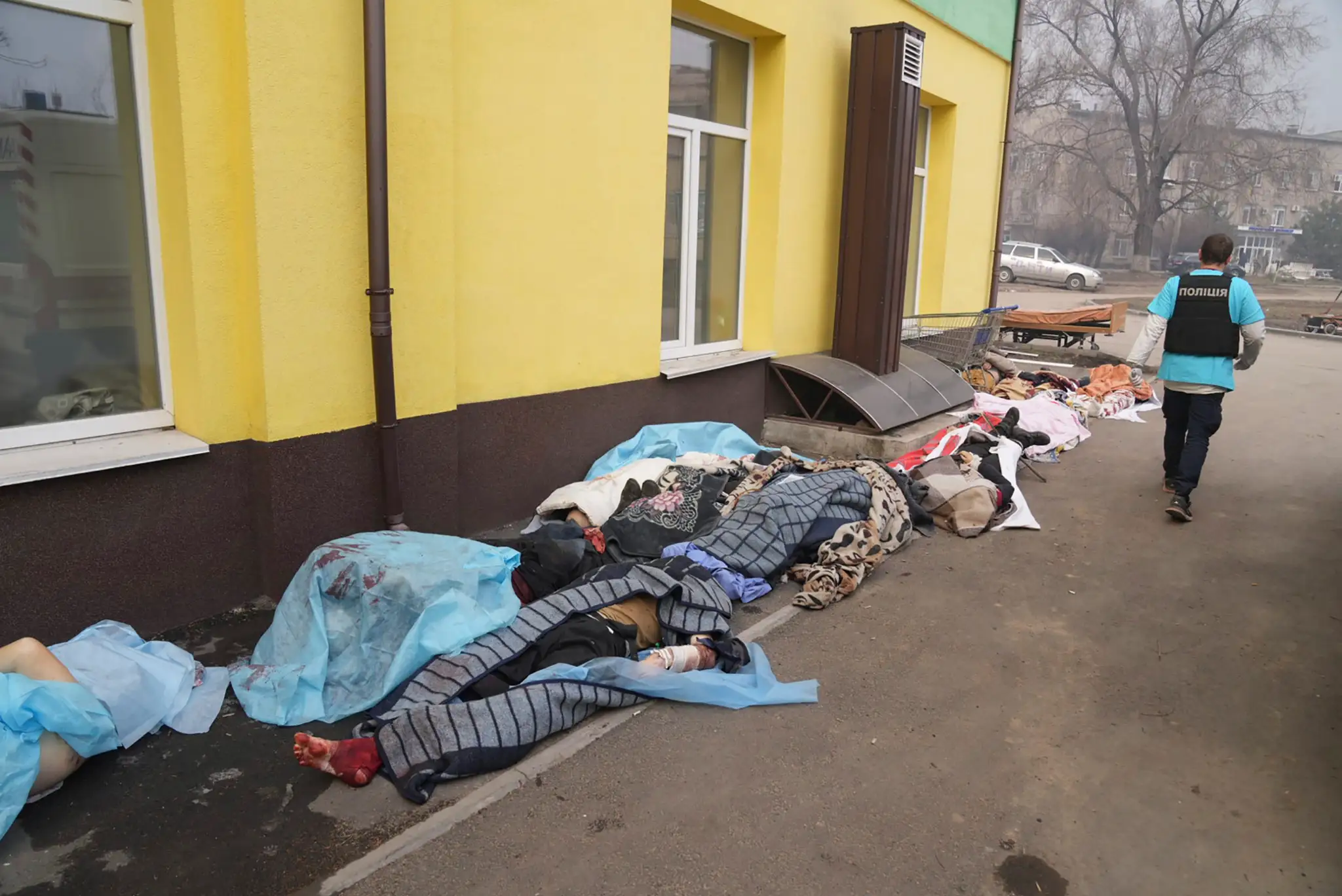 Policajt kráča popri mŕtvych telách pred nemocnicou v Mariupole.