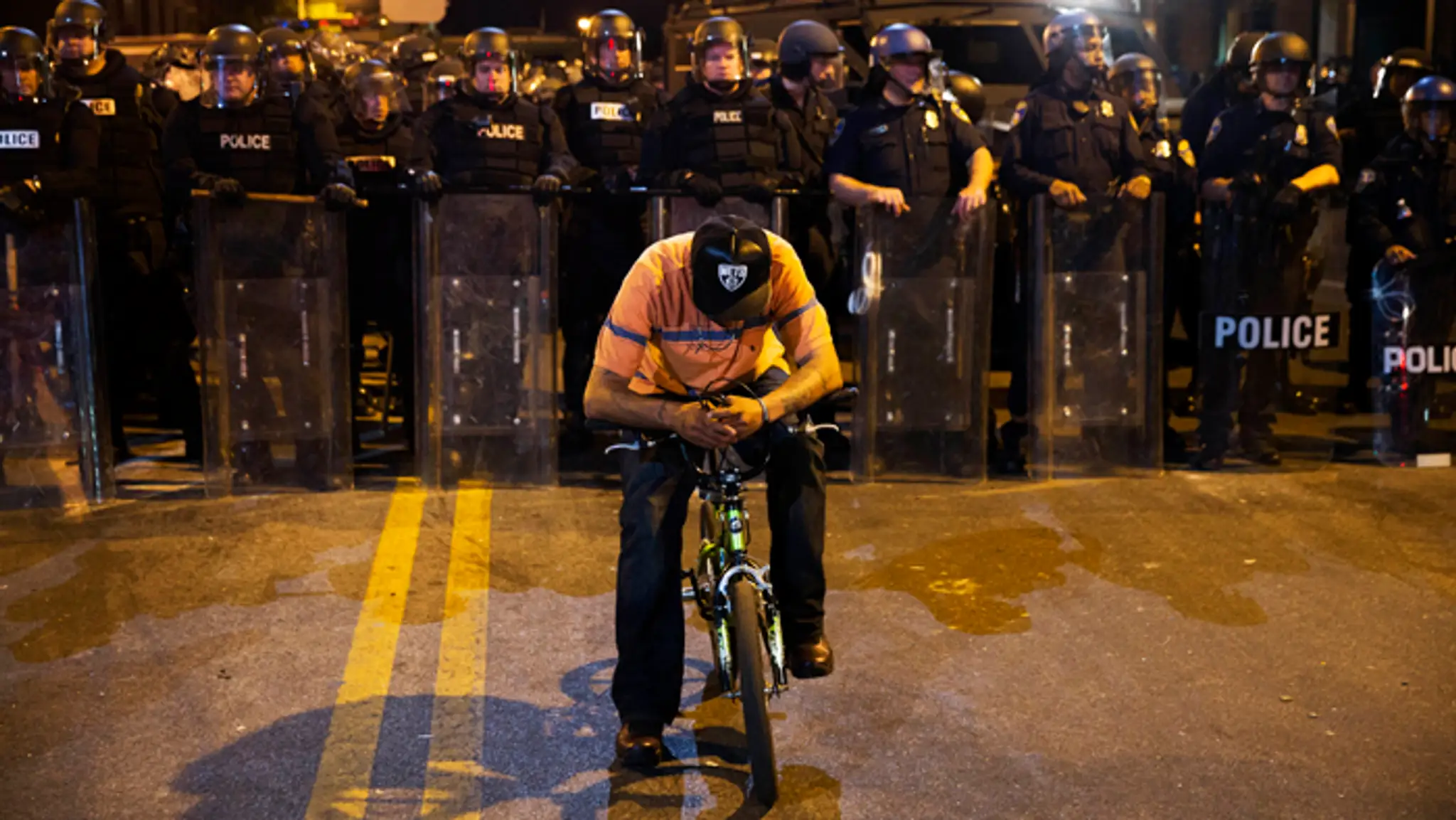 Muž sedí na bicykli pred policajnou líniou počas zákazu vychádzania v Baltimore.