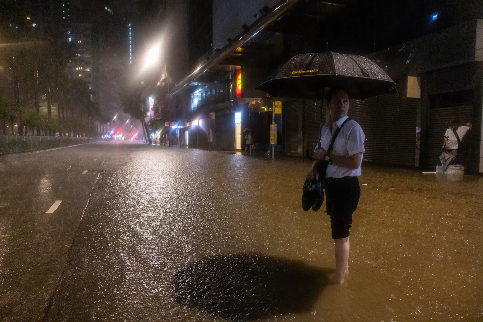 Muž drží v rukách topánky a čaká na autobus na zaplavenej ulici po rekordných dažďových zrážkach v Hongkongu.