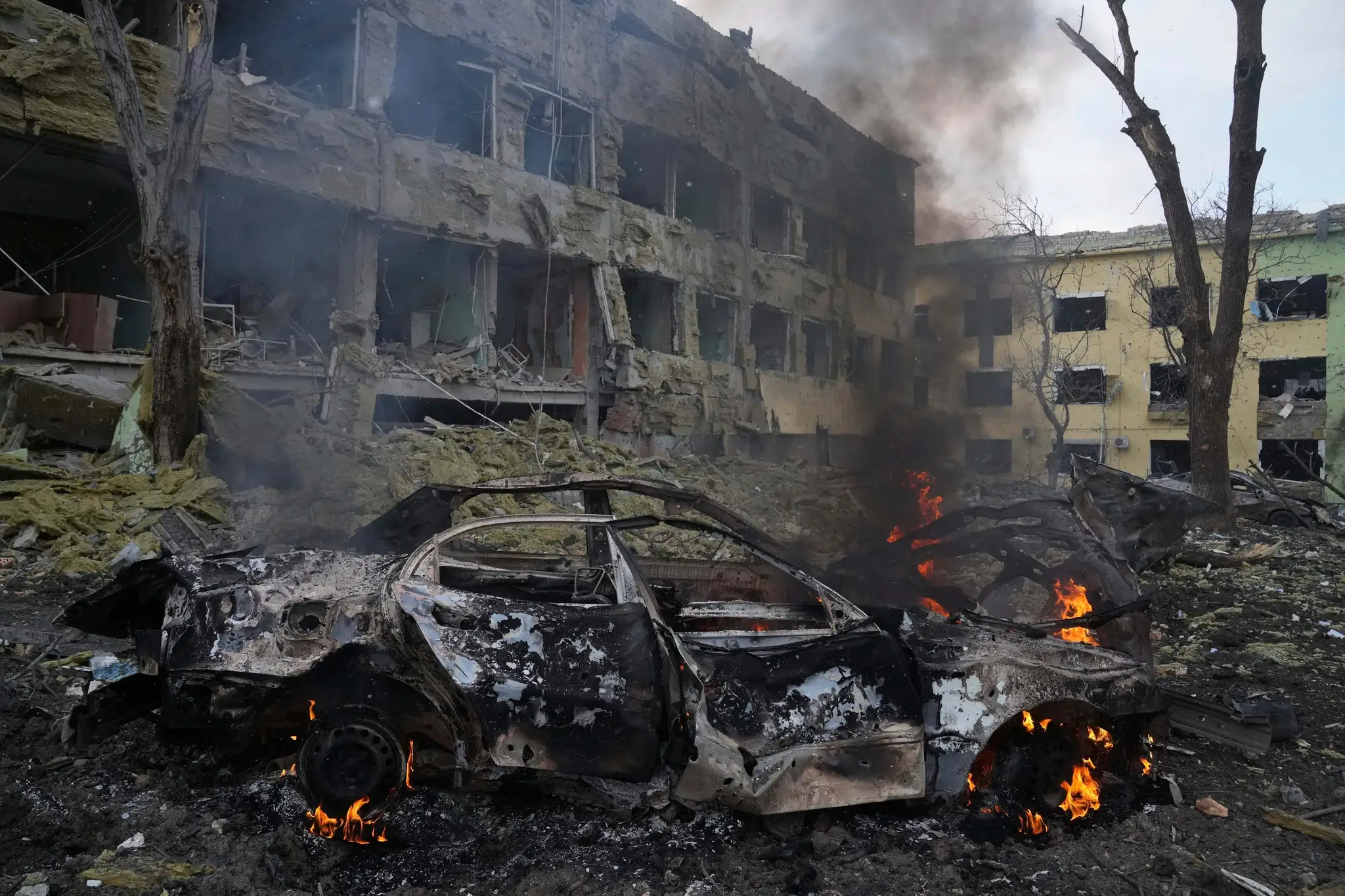 Horiace auto pred bombami zničenou pôrodnicou v Mariupole.