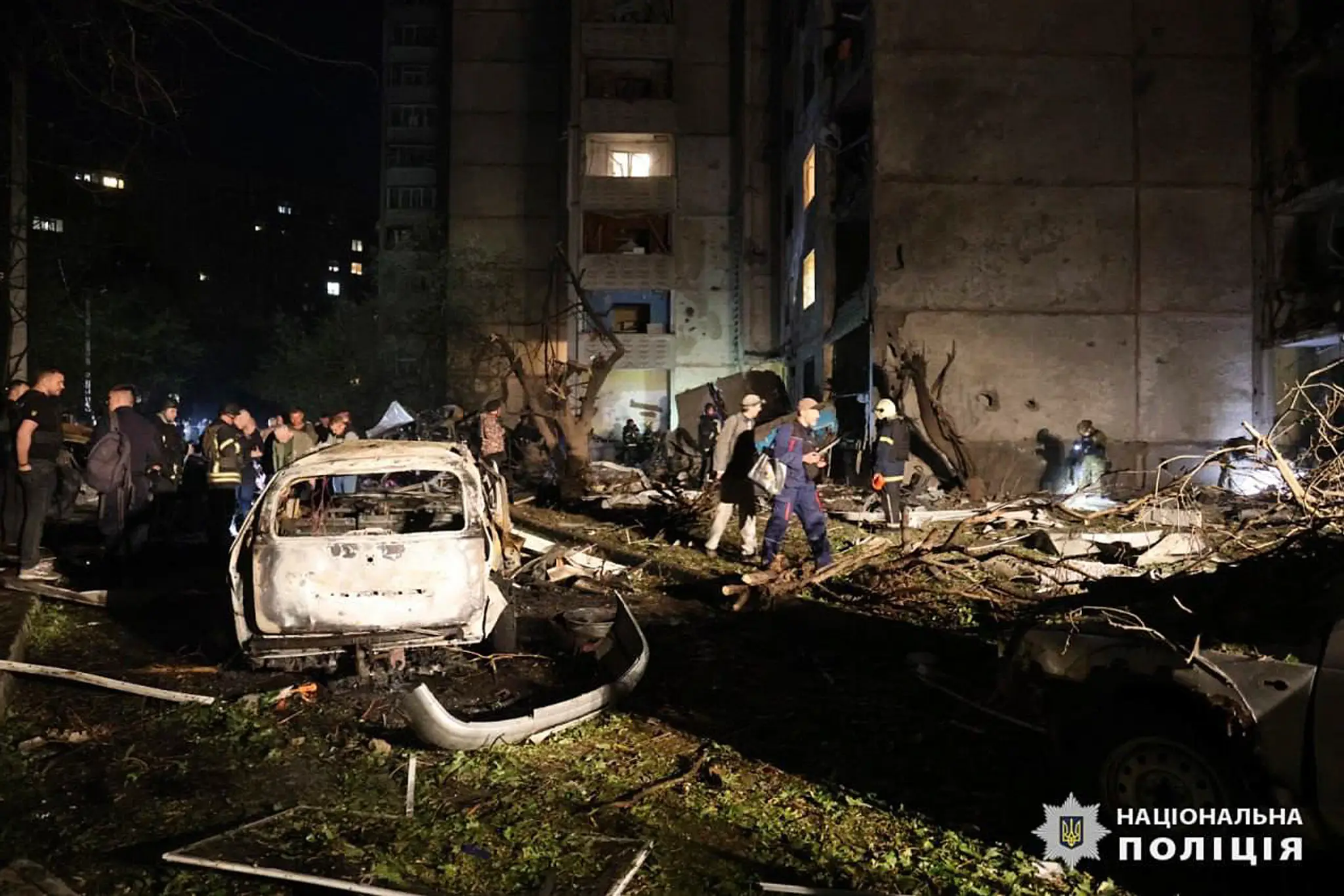 Ľudia stoja pri vrakoch áut po bombovom útoku ruskej armády na obytnú budovu v ukrajinskom meste Charkov.