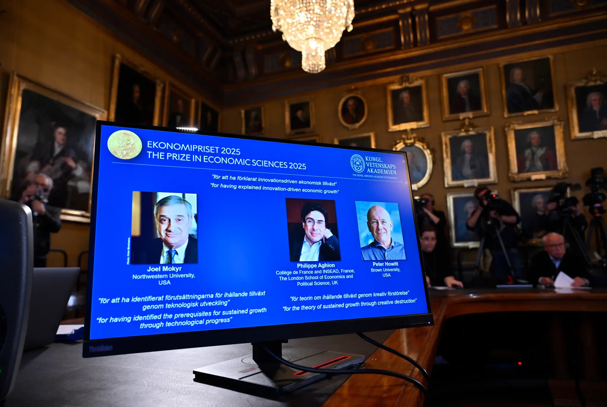 Na veľkoplošnej obrazovke sú hore zľava Joel Mokyr, Philippe Aghion a Peter Howitt, ktorým udelili Nobelovu cenu za ekonómiu za rok 2025.