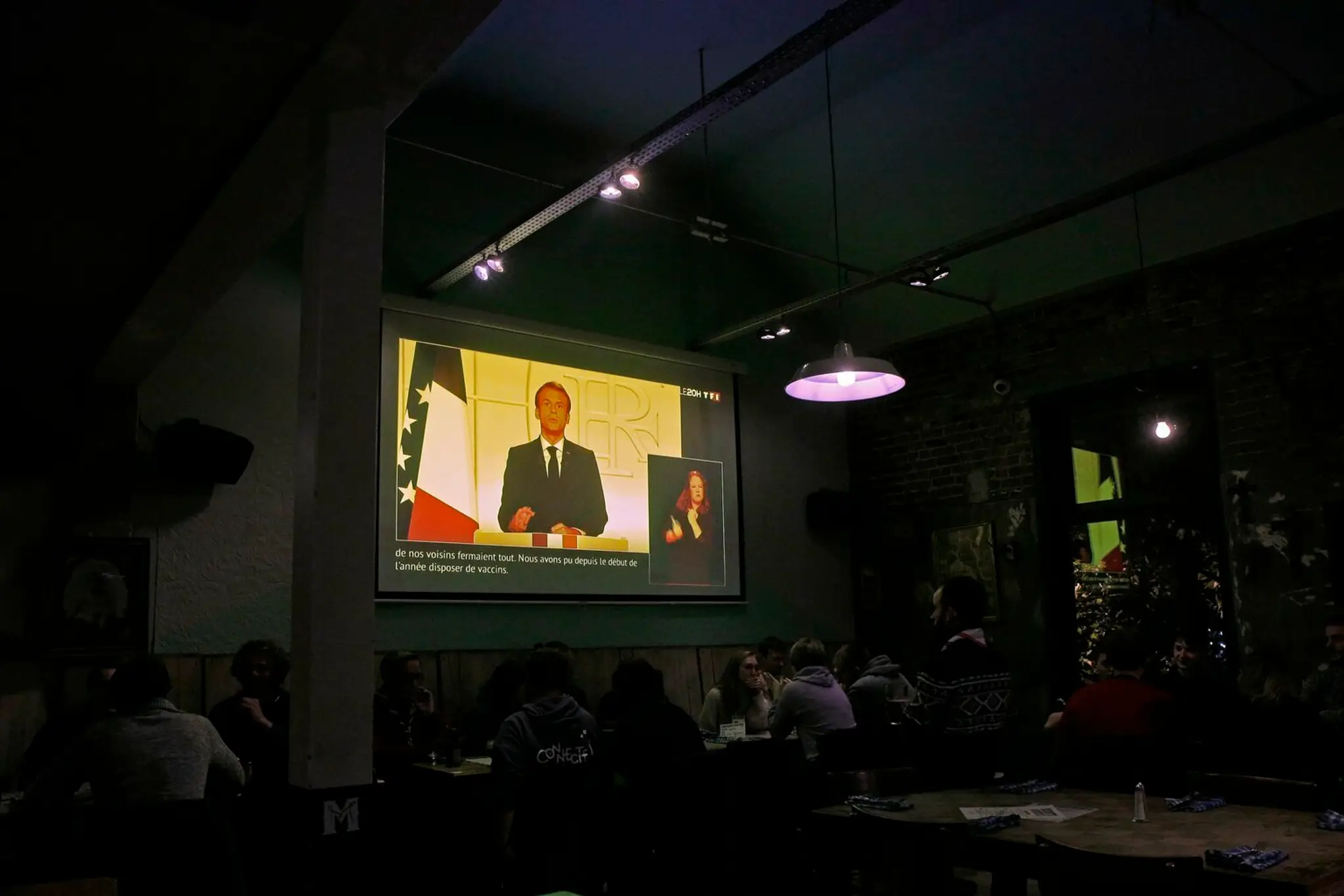 Ľudia sa zabávajú v bare a sledujú televíziu, kde francúzsky prezident Emmanuel Macron vystupuje s televíznym príhovorom.