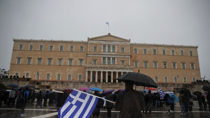 Muž stojaci s gréckou vlajkou pred tamojším parlamentom.