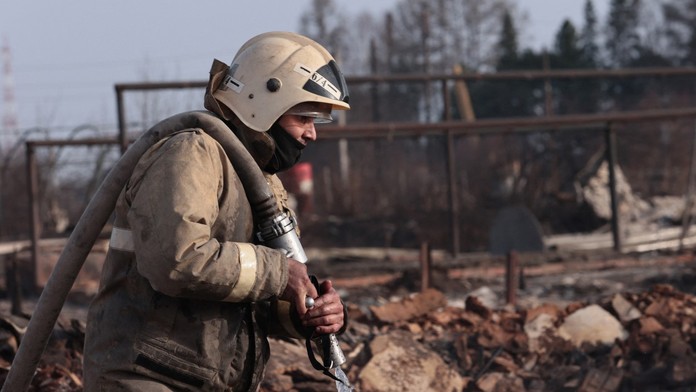 Hasič, ktorý má v ruke hadicu a okolo neho je zhorená dedina.