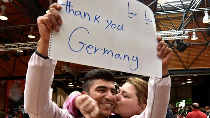 Utečenec z Afganistanu dostáva bozk na líce, keď ukazuje vytrhnutú stránku zo zošita s nápisom "Ďakujeme Nemecko" počas čakania 