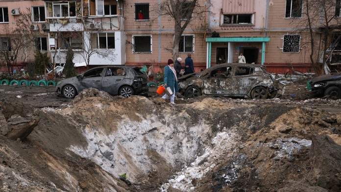 Ľudia prechádzajú okolo krátera a zničených áut pri obytnej budove po ruskom dronovom útoku v Záporoží.