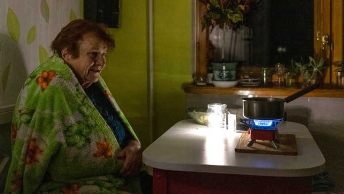 Staršia žena sedí zabalená v  deke a varí jedlo na plynovom horáku počas výpadku elektriny v Kyjeve.