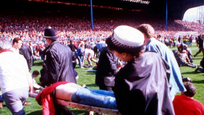 Policajti a fanúšikovia odnášajú zraneného futbalového fanúšika.