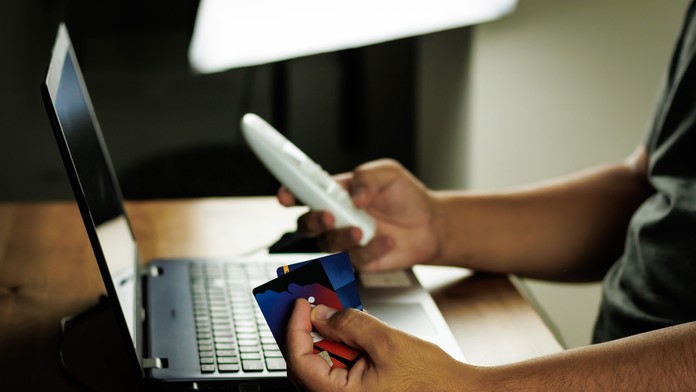 Muž sedí za počítačom, v ruke drží telefón a kreditnú kartu. 