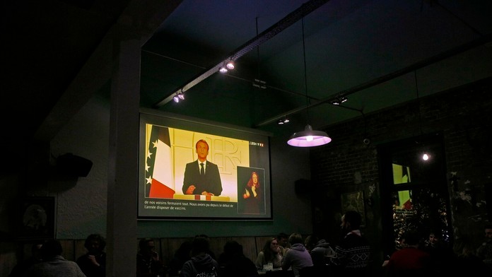 Ľudia sa zabávajú v bare a sledujú televíziu, kde francúzsky prezident Emmanuel Macron vystupuje s televíznym príhovorom.
