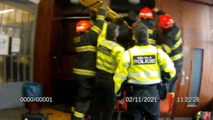 Hasiči s policajtmi sa snažia vyslobodiť seniorku zakliesnenú medzi stenou a výťahom.