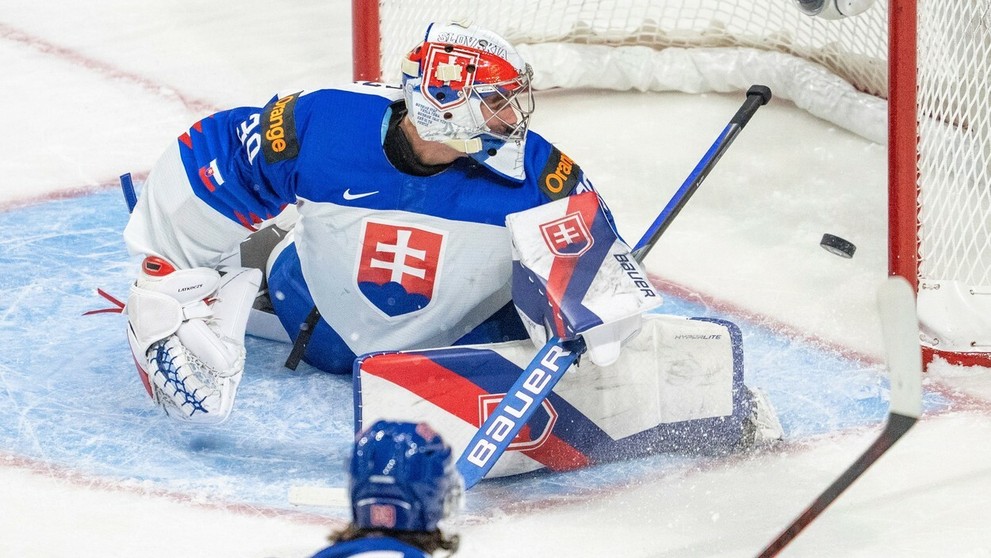Gólman Slovenska Šimon Latkóczy inkasuje v dueli MS juniorov 2022 z hokejky Američana Matta Kniesa.