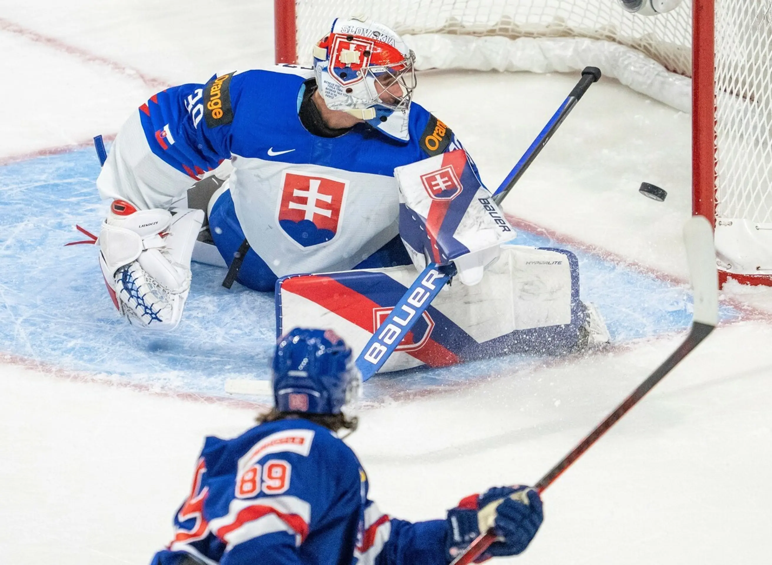 Gólman Slovenska Šimon Latkóczy inkasuje v dueli MS juniorov 2022 z hokejky Američana Matta Kniesa.